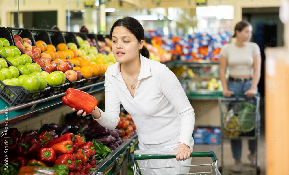 Young woman shopper in casual clothes chooses bell pepper in supermarket