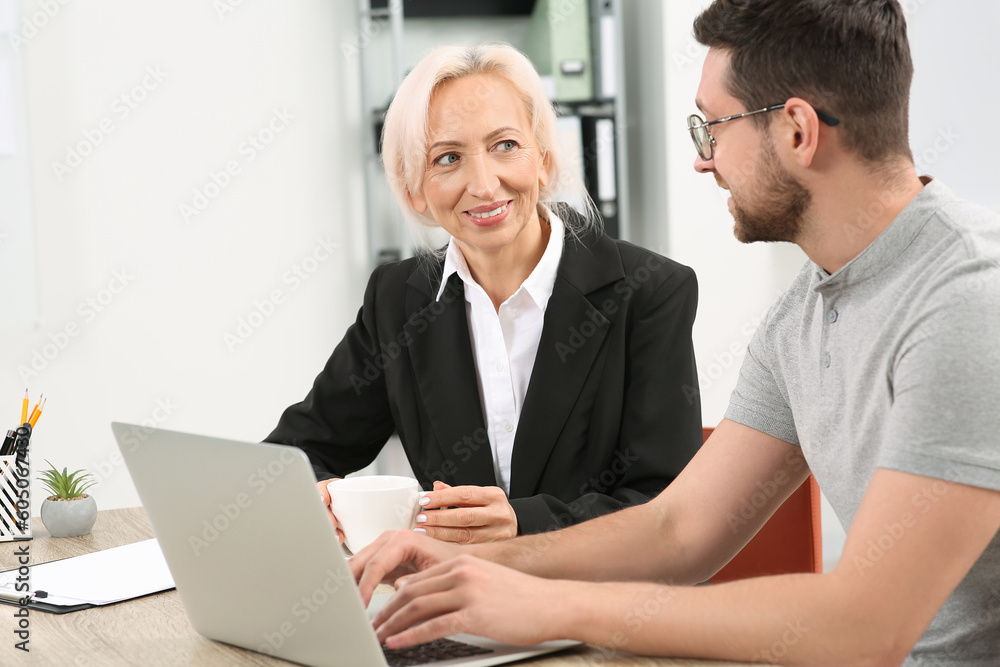 Happy boss with cup of drink and employee discussing work issues at wooden table in office Happy boss with cup of drink and employee discussing work issues at wooden table in office