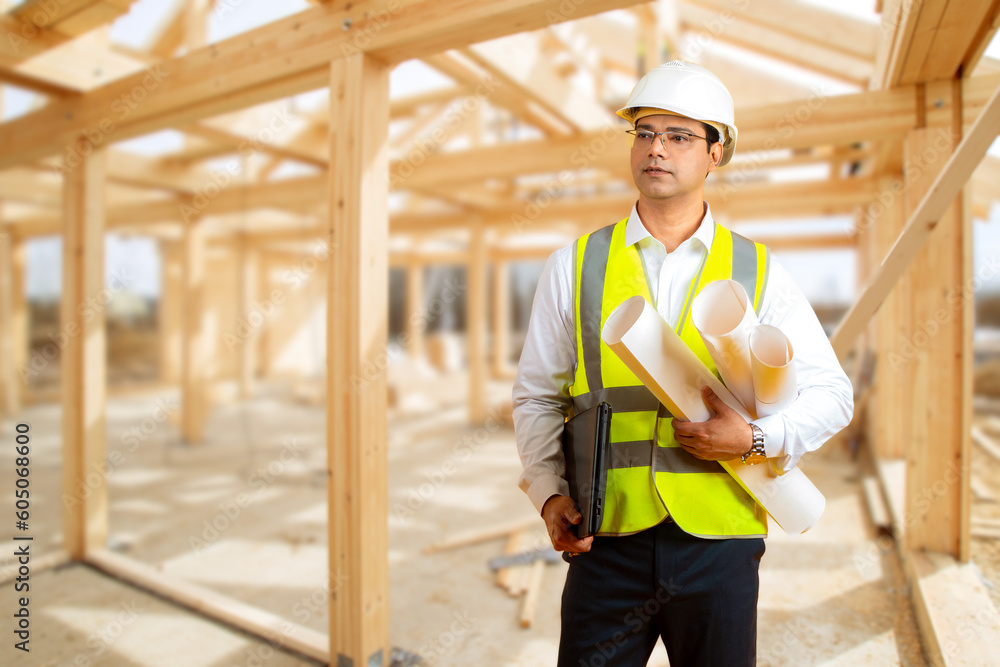 Man builder. Guy with working documentation. Builder near frame of wooden house. Indian man at construction site. Architectural contractor. Construction worker looks ahead thoughtfully Man builder. Guy with working documentation. Builder near frame of wooden house. Indian man at construction site. Architectural contractor. Construction worker looks ahead thoughtfully