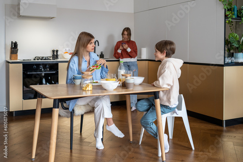 Breakfast on kitchen, mom with son sit at table, talk and eat, daughter chatting in phone in social media. Happy everyday life of family with teens. Healthy nutrition, motherhood, child care concept.