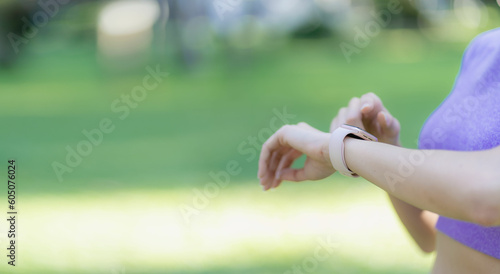 Asian woman standing and looking at her watch during exercise in park or garden with green plants, fitness exercise concept