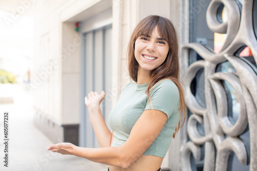pretty young woman feeling happy and cheerful, smiling and welcoming you, inviting you in with a friendly gesture