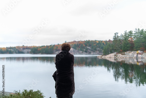 A woman wrapped in a blanket facing and looking at the lake with fall colours in the back