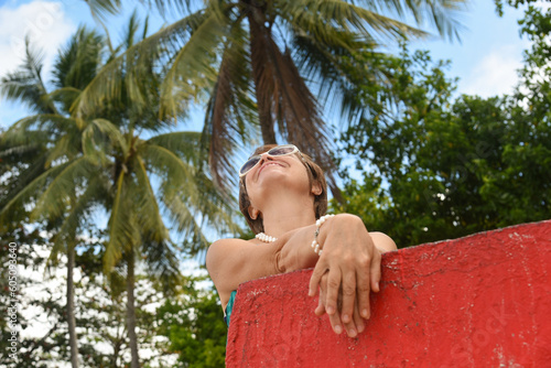 Portrait of senior woman 50-60 age looking up to the sky. Tourist standing near by the red wall on the beach. Concept - travel, retirement, active elderly people.