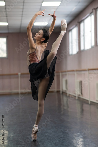 Graceful Asian ballerina in a beige bodysuit and black skirt is rehearsing in a dance class. 
