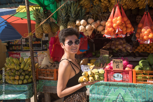Young female buys fruits on street market. Portrait of beautiful girl holding fresh bananas. Woman with short black hair wears sunglasses having fun in vacation. Concept - tourism, travel, vegetarian.