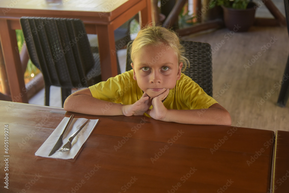 An american blonde boy waiting for a dinner in restaurant. Portrait of child with long blonde hair and a yellow T-shirt sitting in a cafe. Bad appetite and digestive problems on a diet An american blonde boy waiting for a dinner in restaurant. Portrait of child with long blonde hair and a yellow T-shirt sitting in a cafe. Bad appetite and digestive problems on a diet