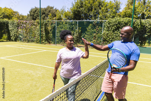 Happy senior african american couple with tennis rackets high fiving over net on sunny tennis court