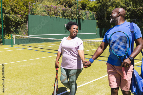 Happy senior african american couple with rackets walking holding hands on sunny grass tennis court
