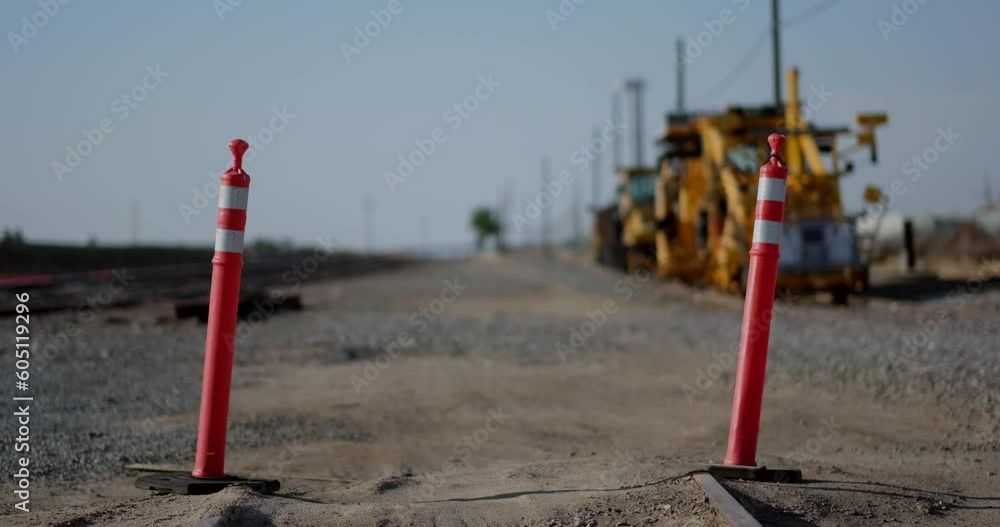 The camera tilts up from the end of disused train tracks to focus on two orange round delineator posts with high-intensity yellow bands. In the defocused background, heavy machinery is visible. The camera tilts up from the end of disused train tracks to focus on two orange round delineator posts with high-intensity yellow bands. In the defocused background, heavy machinery is visible.