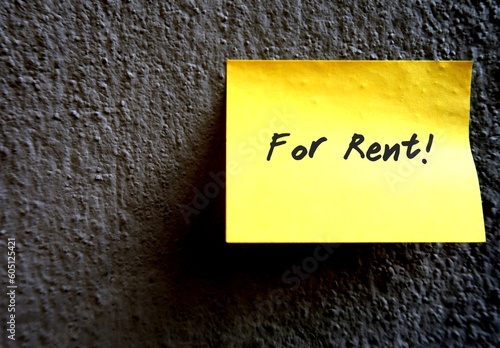 A yellow note pad on the grey rough textured cement wall, with a message FOR RENT, concept of rental business