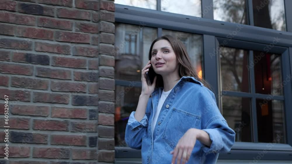 Beautiful young woman talking on phone while walking in city, low angle view. Camera moving left, slow motion Beautiful young woman talking on phone while walking in city, low angle view. Camera moving left, slow motion