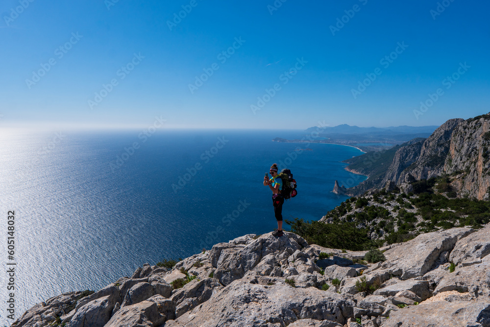Attractive blond young woman looking at the sea while standing on a cliff with a red backpack. Shot in Capo Mannu, Sardinia, Italy.