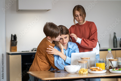 Mom having problems at work working online. Teen children supported her in stressful moment. Boy and girl hug mother working on laptop sitting at table on kitchen at home. Combining family and career.