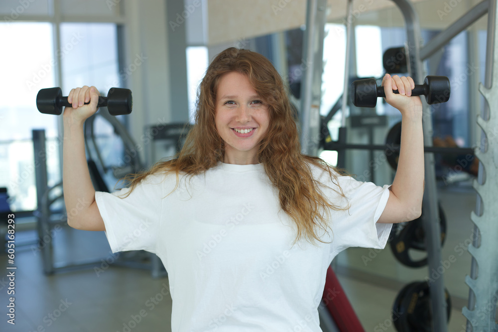 Fototapeta premium Portrait of happy young fit sportive woman training in the gym looking at camera and smile in white T-shirt. Fitness, sport and healthy lifestyle concept.