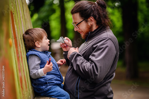 Tall long dark haired man with ponytail and glasses stands near wooden fence with little toddler boy