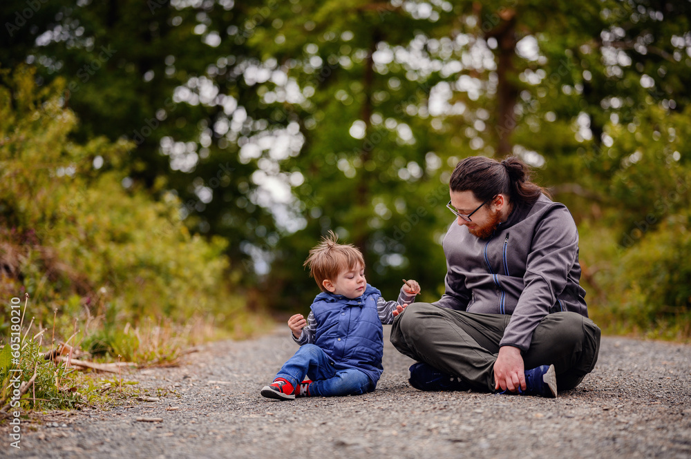 Fototapeta premium Young long hair bearded man in glasses sitting with little toddler boy on road playing with stones