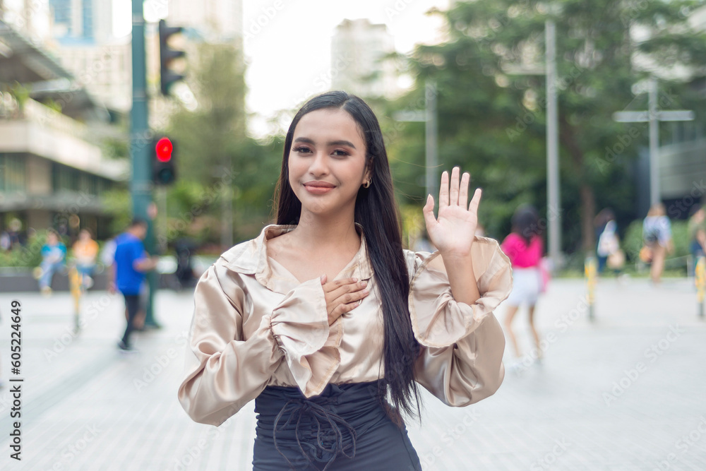 Fototapeta premium A pretty woman standing outside wearing a light tan silk blouse in an oath taking pose while smiling at the camera. Trees, building, people and traffic light are shown in the background.