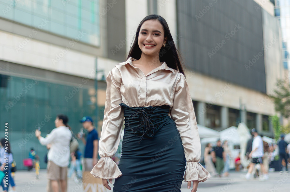 A pretty asian woman standing confidently outside with poise and wearing a light tan silk blouse and a high waist fitted black skirt. A building, people and tents in the background