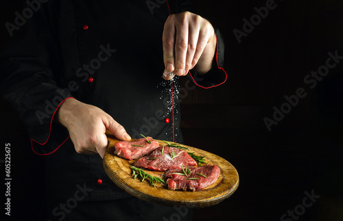 A professional butcher or chef salts raw beef steaks before a barbecue. Preparation for roasting veal meat with rosemary and spices. Black space for recipe or restaurant menu