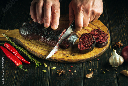 The chef cuts blood sausage on kitchen cutting board with knife in his hand. The concept of cooking a national dish with spices and peppers at home in kitchen. Bull blood sausage according to  photo