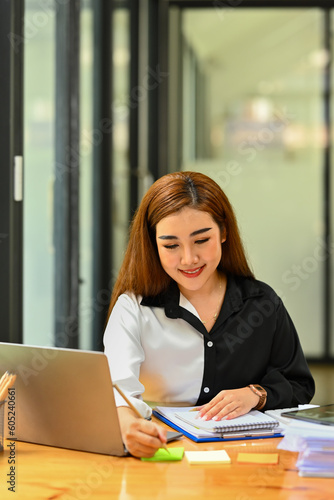 Smiling female employee sitting in front of laptop and writing on sticky notes © Prathankarnpap