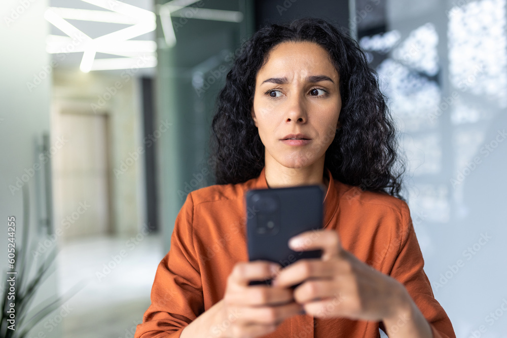 Upset thinking business woman close up inside office, young hispanic woman reading bad news on phone online, business woman at workplace holding smartphone typing text message. Upset thinking business woman close up inside office, young hispanic woman reading bad news on phone online, business woman at workplace holding smartphone typing text message.