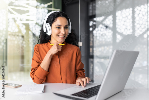 Young curvy businesswoman with headphones listening to online course and audio podcast female worker inside office working with laptop, successful hispanic woman smiling satisfied with work. photo