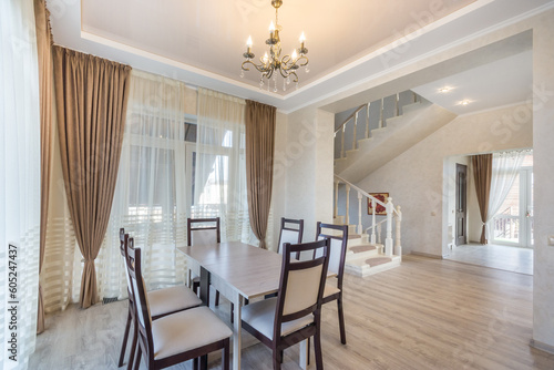 The interior of a country house with a modern design. Elegant chandelier over the dining table, windows with beautiful curtains, stairs to the second floor.