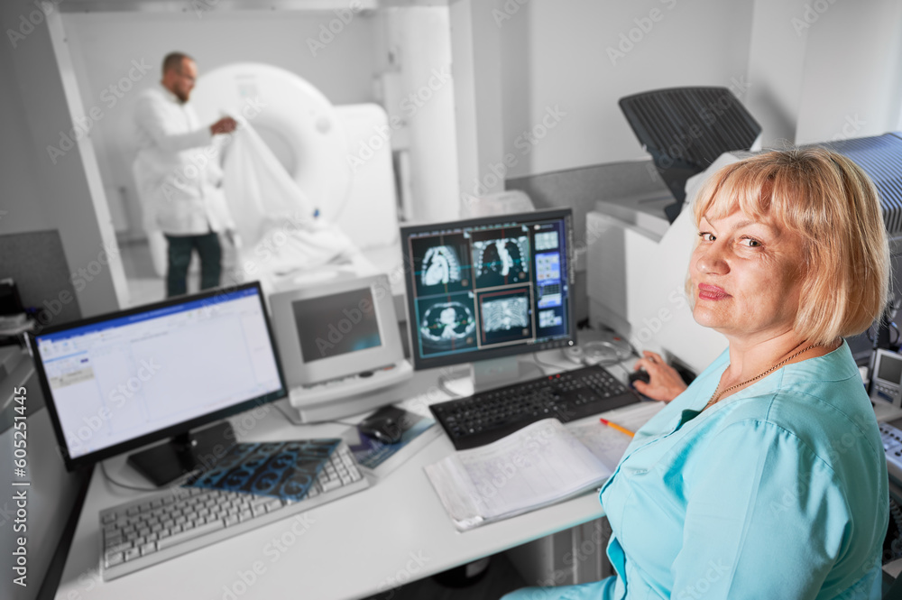 Medical computed tomography or MRI scanner. Portrait of female nurse working at computer, making MRI. Woman wearing blue uniform. Concept of medicine and hospital. Medical computed tomography or MRI scanner. Portrait of female nurse working at computer, making MRI. Woman wearing blue uniform. Concept of medicine and hospital.