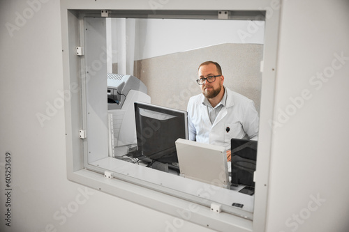 Doctor sitting, working at computer, making MRI. Male specialist wearing glasses and white robe, looking at camera, smiling, Concept of modern diagnostics.