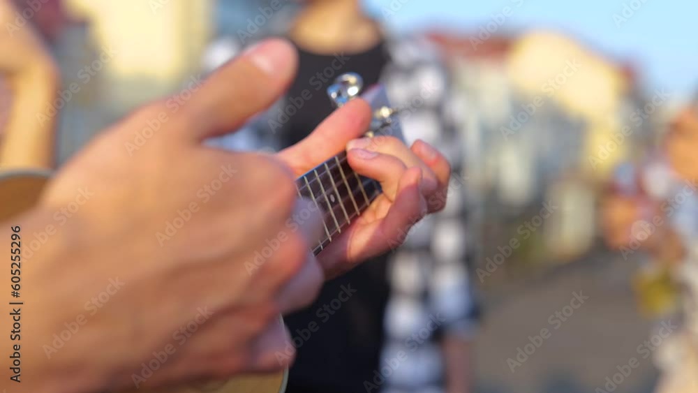 Male hands holding ukulele while playing music with young people dancing on the background. Party on the rooftop outdoors. Lifestyle, friendship, fun concept. Real time Male hands holding ukulele while playing music with young people dancing on the background. Party on the rooftop outdoors. Lifestyle, friendship, fun concept. Real time