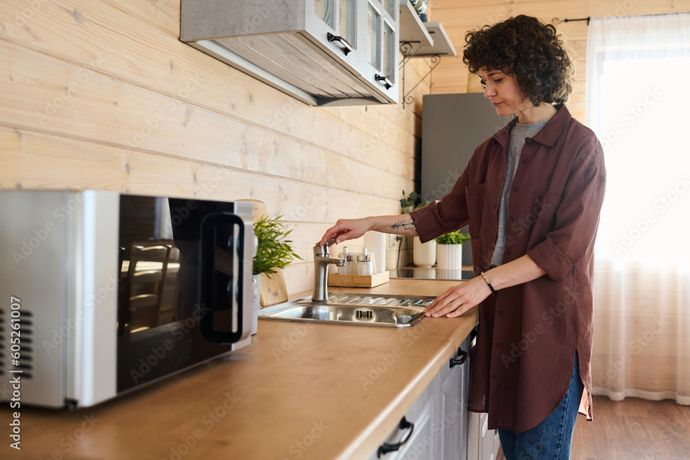 Young woman in casual attire opening facet while standing by wooden kitchen counter with metallic sink in her house with wooden walls Young woman in casual attire opening facet while standing by wooden kitchen counter with metallic sink in her house with wooden walls