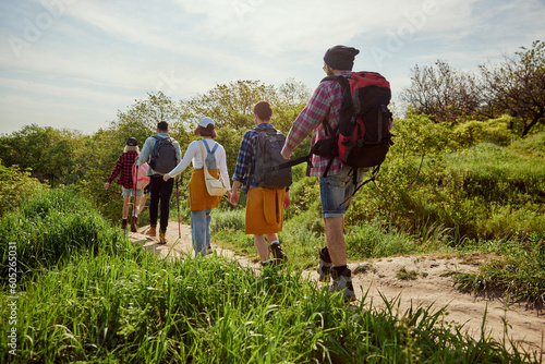 Young people, group of friends in comfortable clothes with backpacks walking in forest, going hiking on warm summer day. Concept of active lifestyle, nature, sport and hobby, friendship