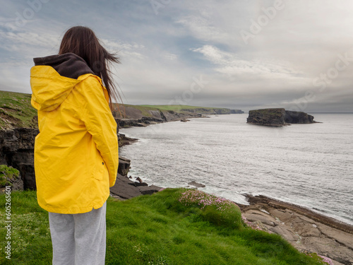 Teenager girl in yellow jacket looking at stunning nature scenery with cliffs and ocean, Kilkee area, county Clare, Ireland. Travel and sightseeing concept. Cloudy sky.