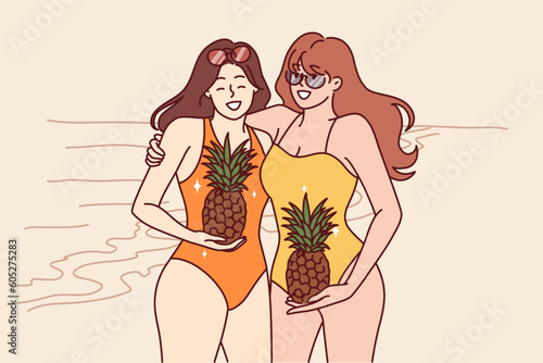 Two women in swimsuits stand on beach with pineapples in hands and smile inviting visit tropical island. Young happy girls in swimsuits spend holidays on sunny beach near sea or ocean