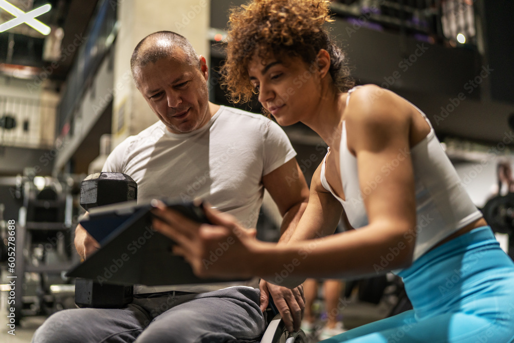 A determined individual in a wheelchair, working out in the gym with the guidance of a dedicated female fitness trainer who using a tablet to show a weekly workout plan. A determined individual in a wheelchair, working out in the gym with the guidance of a dedicated female fitness trainer who using a tablet to show a weekly workout plan.