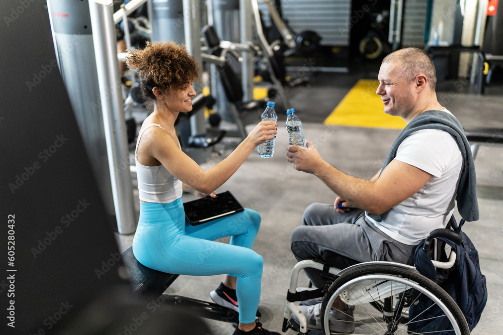 A physically challenged person without obstacles workout at the gym with his female fitness instructor. They're taking a break to refresh and eat energy bars. - obrazy, fototapety, plakaty 