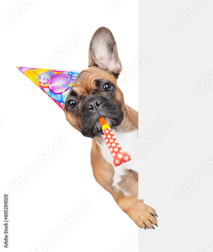 French Bulldog puppy wearing party cap blows into party whistle and looks from behind empty white banner. isolated on white background