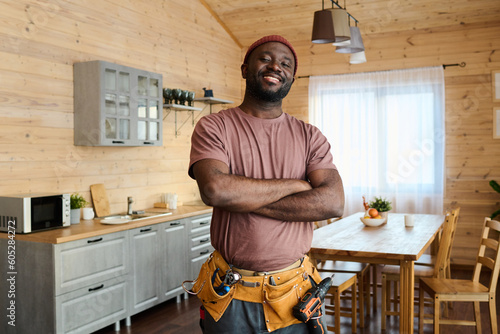 Young cheerful African American repairman or plumber with toolbelt keeping his arms crossed by chest and looking at camera with smile photo