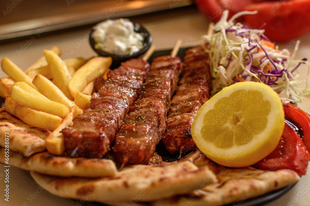 Plate with juicy mouthwatering pork souvlaki roasted on a BBQ grill, served with potato, pita, salad, tomato, lemon and sous in greek tavern. Selective soft focus. Mediterranean cuisine