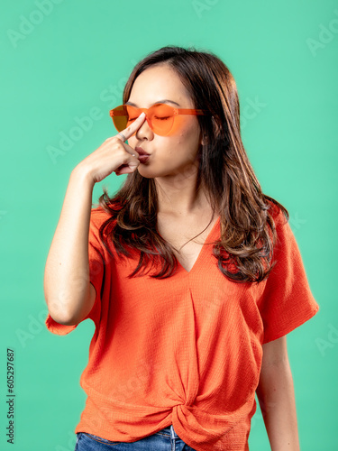 A portrait of an Asian woman wearing an orange shirt and glasses, holding her glasses, isolated on a green background.