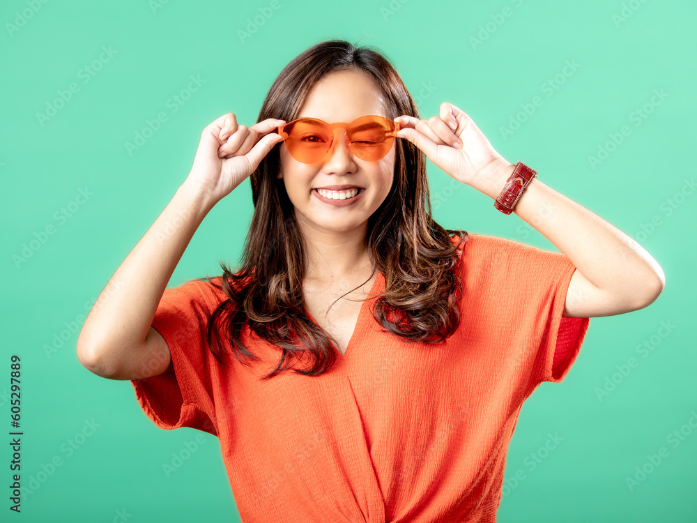 A portrait of an Asian woman wearing an orange shirt and glasses, holding her glasses, isolated on a green background. A portrait of an Asian woman wearing an orange shirt and glasses, holding her glasses, isolated on a green background.