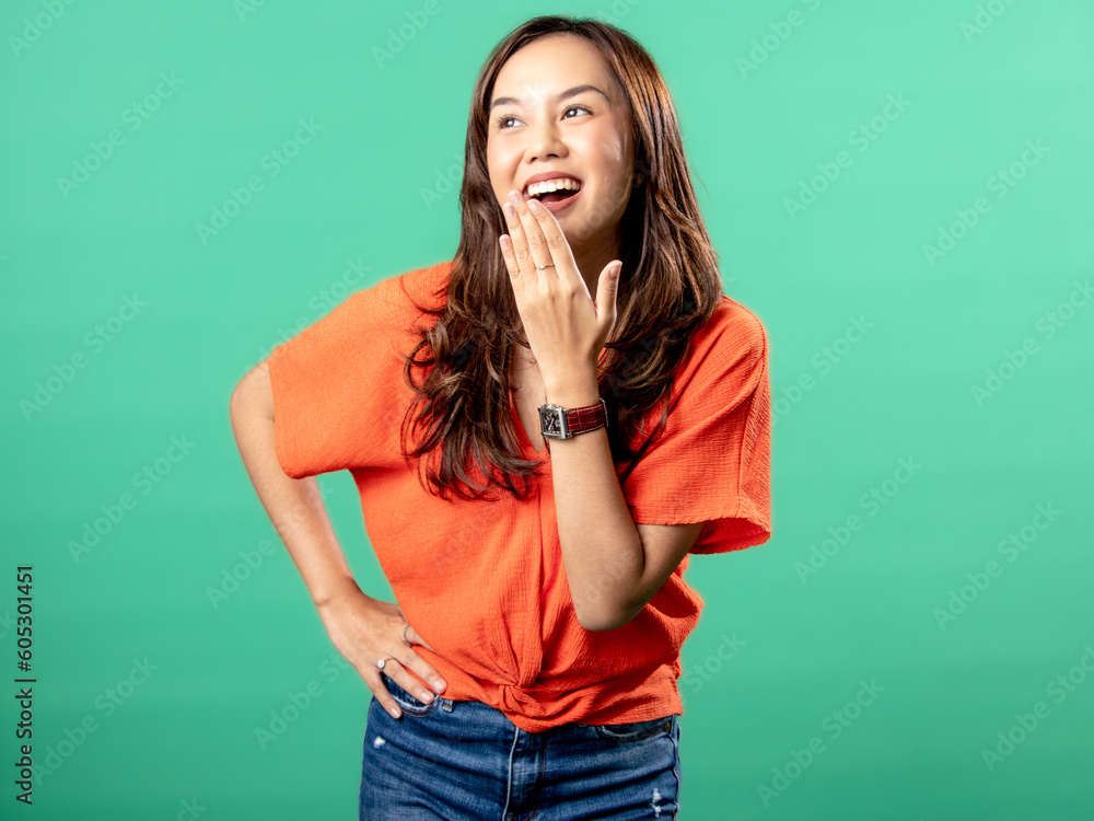 A portrait of an Asian woman wearing an orange shirt, calling out and opening her palm next to her mouth, isolated on a green background. A portrait of an Asian woman wearing an orange shirt, calling out and opening her palm next to her mouth, isolated on a green background.