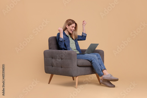 Emotional woman with laptop sitting in armchair on beige background