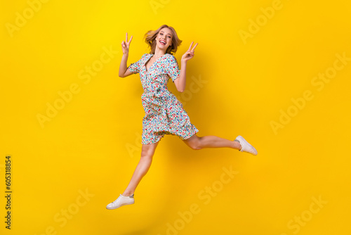 Full length photo of cheerful excited lady wear flower print dress jumping high showing v-signs isolated yellow color background