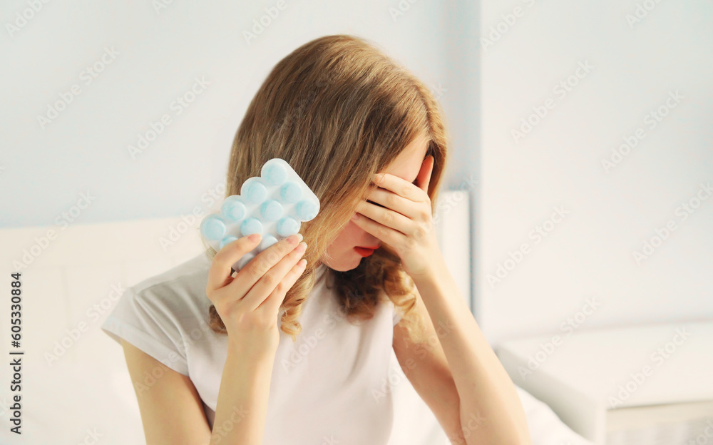 Portrait of sick upset woman holding pills, tired female patient does not want to take medicine closing her eyes with her hands on the bed in bedroom Portrait of sick upset woman holding pills, tired female patient does not want to take medicine closing her eyes with her hands on the bed in bedroom