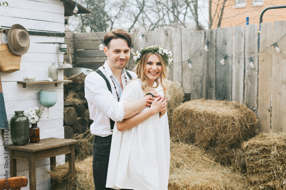 young couple newlyweds bride in short white dress and wreath with veil and groom in hat and jeans with suspenders hugging and having fun at barn of country house young couple newlyweds bride in short white dress and wreath with veil and groom in hat and jeans with suspenders hugging and having fun at barn of country house