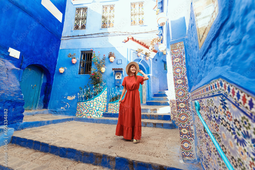 Young woman with red dress visiting the blue city Chefchaouen, Marocco - Happy tourist walking in Moroccan city street - Travel and vacation lifestyle concept - obrazy, fototapety, plakaty 