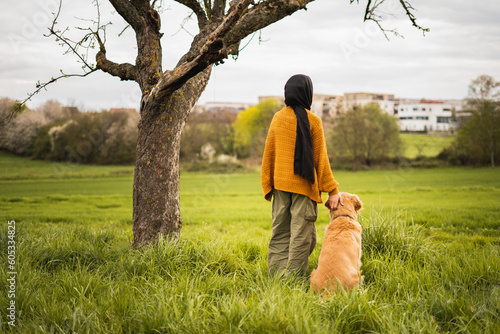 young muslim girl standing with a dog, two best friends under a tree in a park while watching the view on a walk